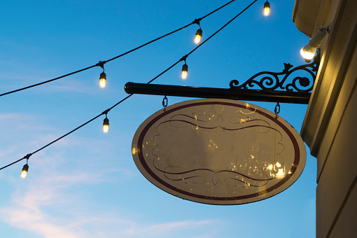 Blank vintage store sign hanging with lights against a blue evening sky
