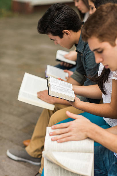 Side view of young people reading Side view of a small group of people sitting in a row and reading each one a book in a vertical medium shot outdoors. bible study group of people small group of people stock pictures, royalty-free photos & images