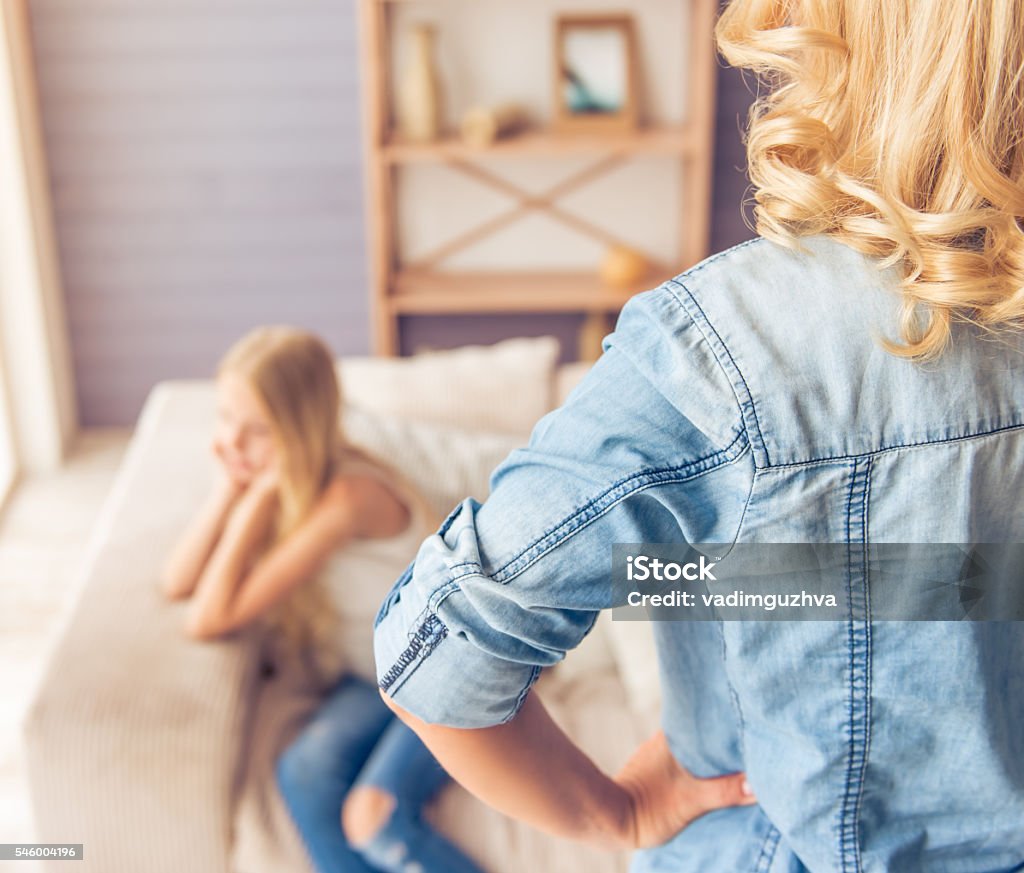 Mom and daughter Back view of woman standing akimbo and looking at her teenage daughter who is sitting sadly on sofa in the background Child Stock Photo