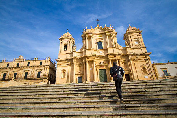 Noto, Sicily: Man Walking up Steps toward Cathedral A man walking up the steps to the baroque cathedral in Noto, Sicily, which dates from the 1700s. The building operates exclusively as a house of worship, and entry is open and free to all. Noto is a World Heritage site. Copy space in the blue sky. noto sicily stock pictures, royalty-free photos & images