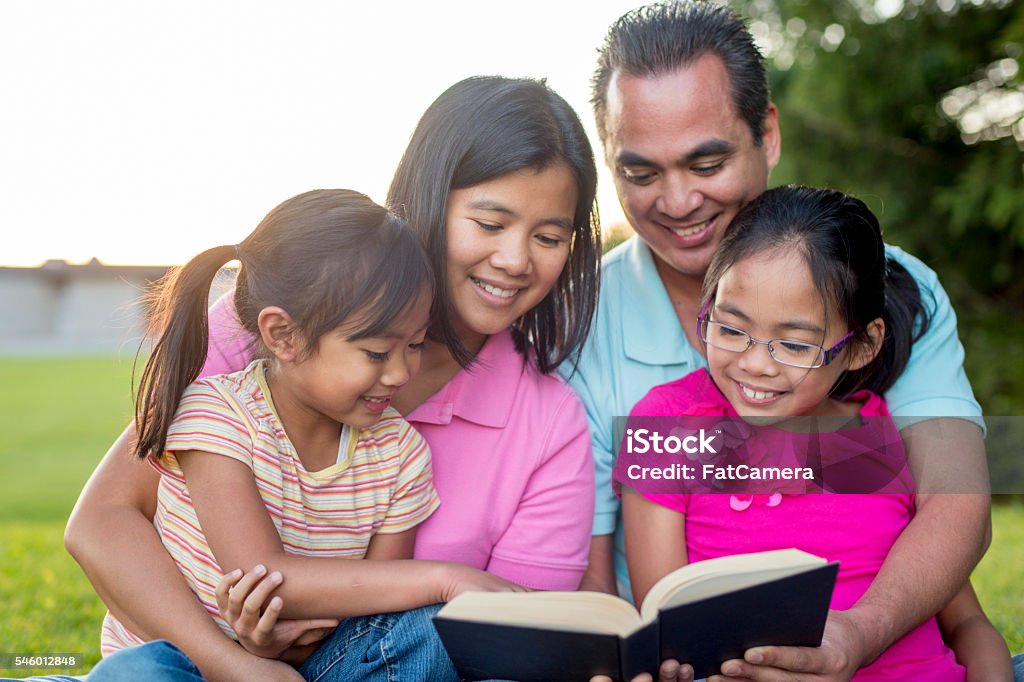 Reading the Bible as a Family A family of four is sitting in the grass at the park and are reading the Bible together. Bible Stock Photo