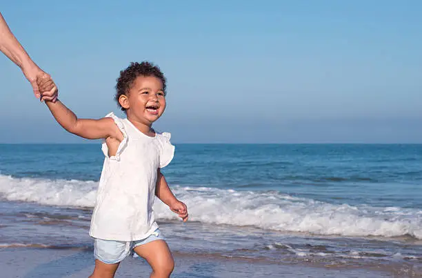 2.5 years old girl holding her grandmother's hand, jogging on the sandy beach, looking away with a big excited smile.