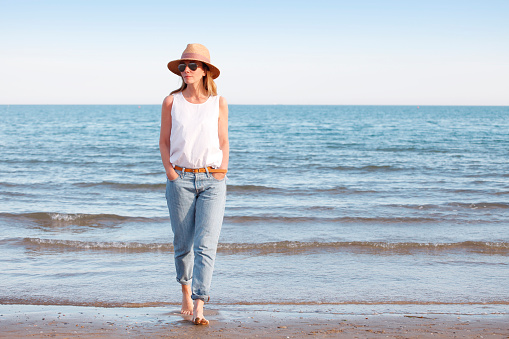 Full length portrait of an attractive mature woman walking on the beach barefoot. Beautiful female in straw hat and sunglasses wearing casual clothes.