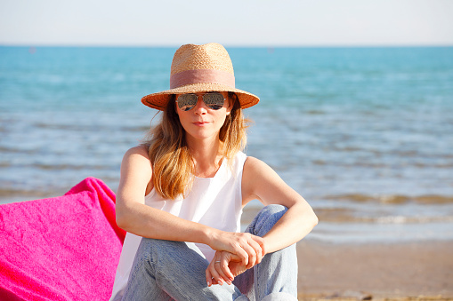 Close-up of happy mature woman enjoying summer vacation while sitting on sandy beach and relaxing.