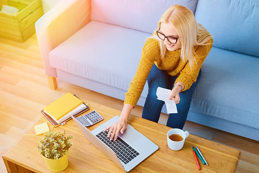 Happy  young woman paying bills on laptop sitting on sofa