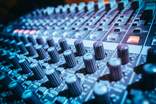 Horizontal composition color photography of close-up in selective focus of DJ's sound mixer with faders illuminated by blue stage light, sound mixer button in concert.