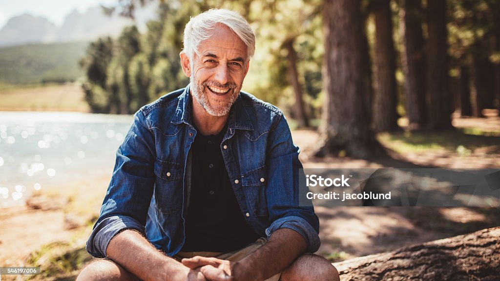 Happy mature man sitting near a lake Portrait of happy mature man sitting near a lake looking at camera and smiling. Senior caucasian man sitting on a log by the lake on a summer day. Men Stock Photo