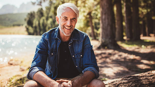 Happy mature man sitting near a lake Portrait of happy mature man sitting near a lake looking at camera and smiling. Senior caucasian man sitting on a log by the lake on a summer day. mature men stock pictures, royalty-free photos & images