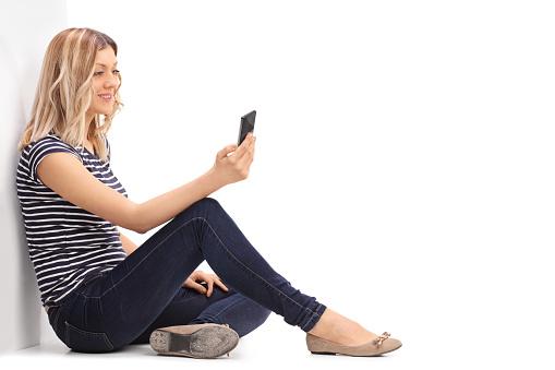 Young woman sitting on the floor and reading a text message on her phone isolated on white background
