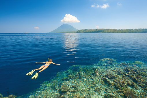 Young woman in swimsuit snorkeling in blue and transparent tropical sea on a background volcano Manado Tua.  North Sulawesi, Indonesia.