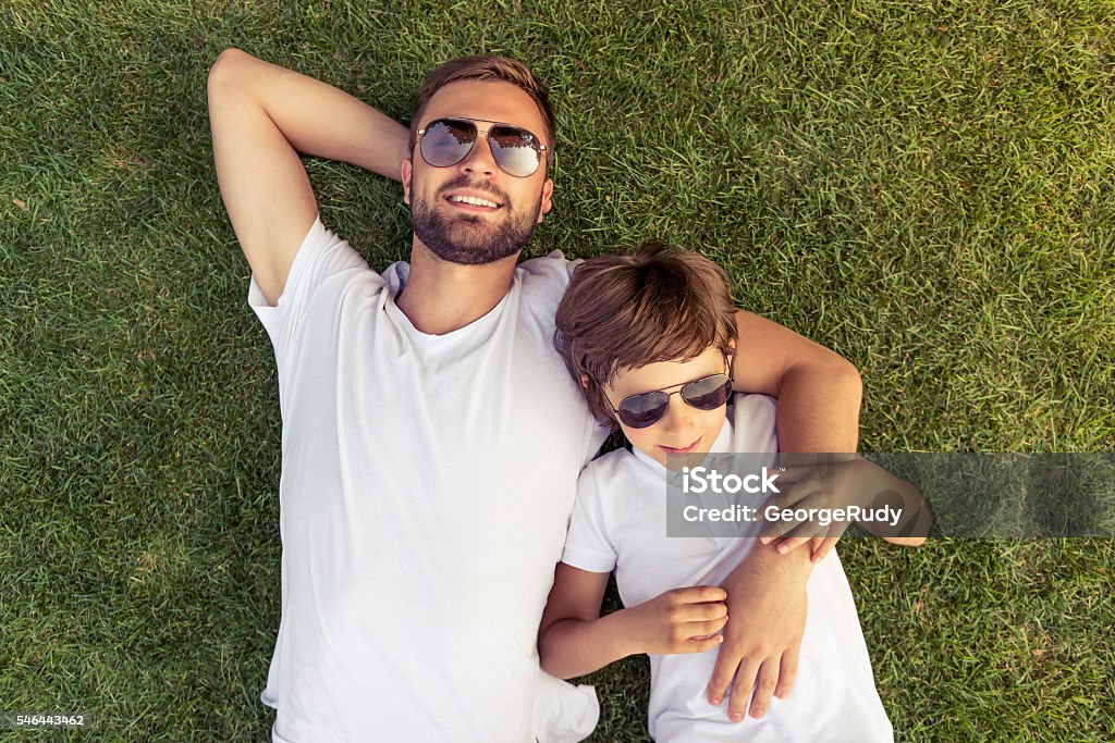 Dad and son resting outdoors Top view of cute little boy and his handsome young dad in white T-shirts and sun glasses smiling while on the grass Father Stock Photo