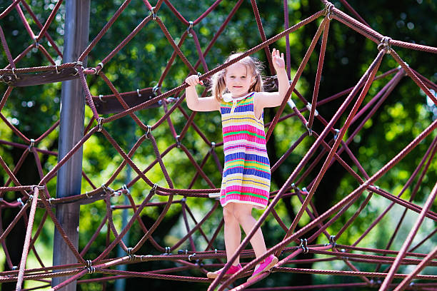 Active little child playing on climbing net and jumping on trampoline at school yard playground. Kids play and climb outdoors on sunny summer day. Cute girl on nest swing at preschool sport center.