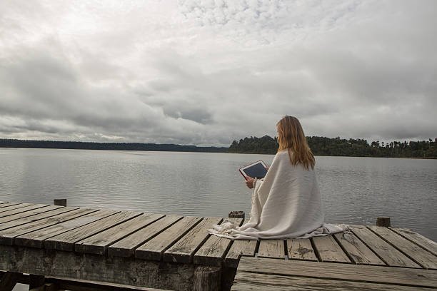 istock Woman sits on jetty above lake, uses digital tablet 546451826
