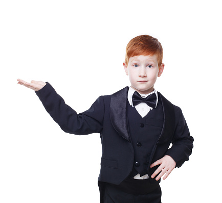 Little cute funny redhead boy in tailcoat tuxedo show something pointing at nowhere. Portrait of well-dressed child in bow tie isolated on white background