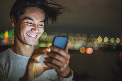 Close-up of a young Japanese man texting outdoors at night, with city lights at the background and copy space.