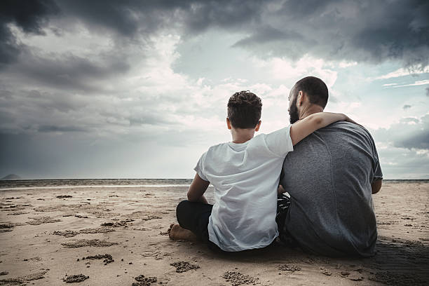 Father and son looking at sunset sitting on the beach. Stock photo.