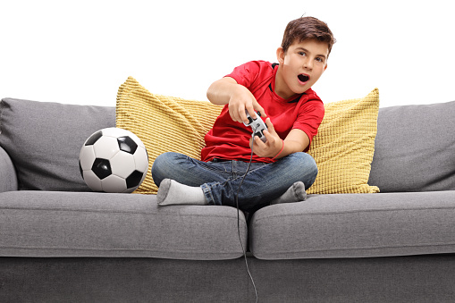 Little boy playing football video game seated on a sofa isolated on white background