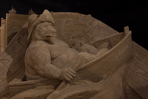Sand sculpture of a bear with tattoo and hat looking annoyed while rowing a boat. This sand sculpture is made by Bouke Atema and photograped by Bouke Atema.