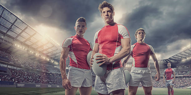 Heroic Rugby Players Standing With Ball On Pitch In Stadium A low angle portrait of four heroic male rugby players wearing unbranded red and white rugby kit. The teammates standing together, looking into the distance, and holding rugby balls on an outdoor rugby pitch in a generic floodlit stadium full of spectators, under a dramatic stormy evening sky at sunset.  rugby team stock pictures, royalty-free photos & images