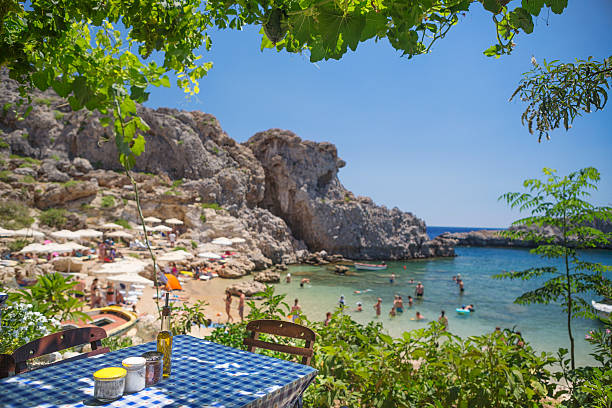 tisch einer traditionellen griechischen taverne mit strandhintergrund - lindos stock-fotos und bilder