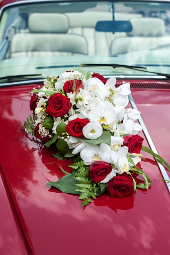 Vertical close-up image of beautiful bunch of roses with orchids attached on luxury red car, ready to transport on wedding ceremony. Elegant floral wedding decoration.