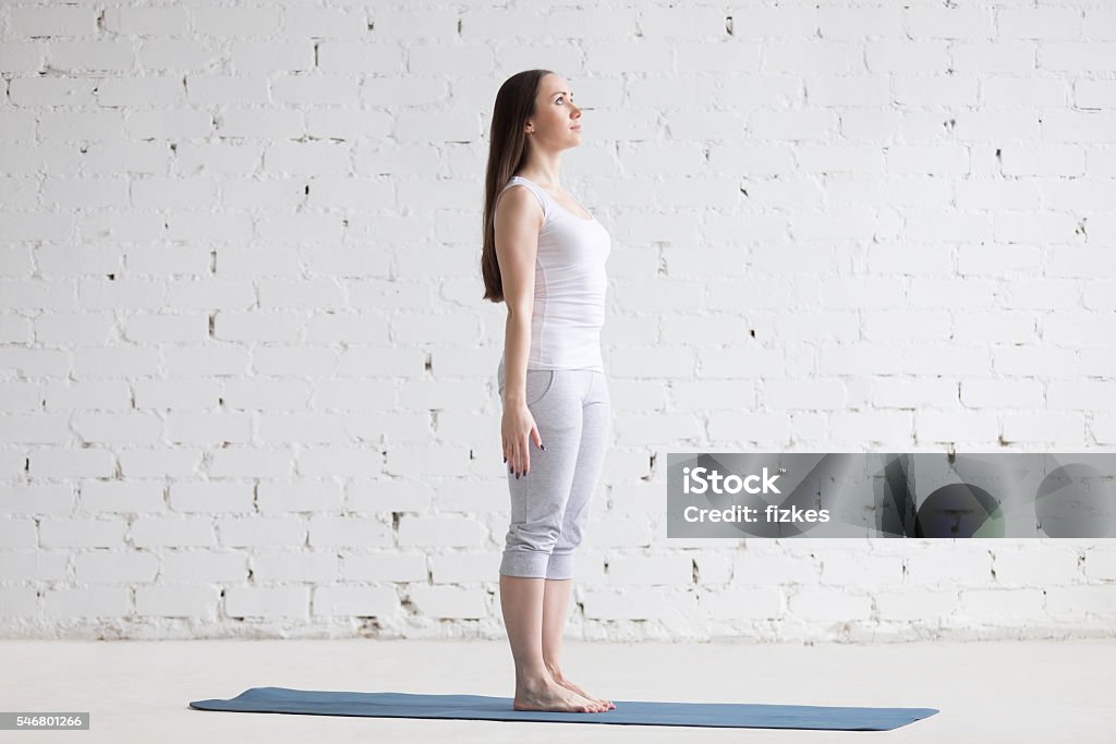 Beautiful sporty young woman doing Tadasana pose in white loft Attractive happy young woman working out indoors. Side view portrait of beautiful model doing yoga exercise on blue mat. Standing in Tadasana, Mountain Pose. Full length Good Posture Stock Photo
