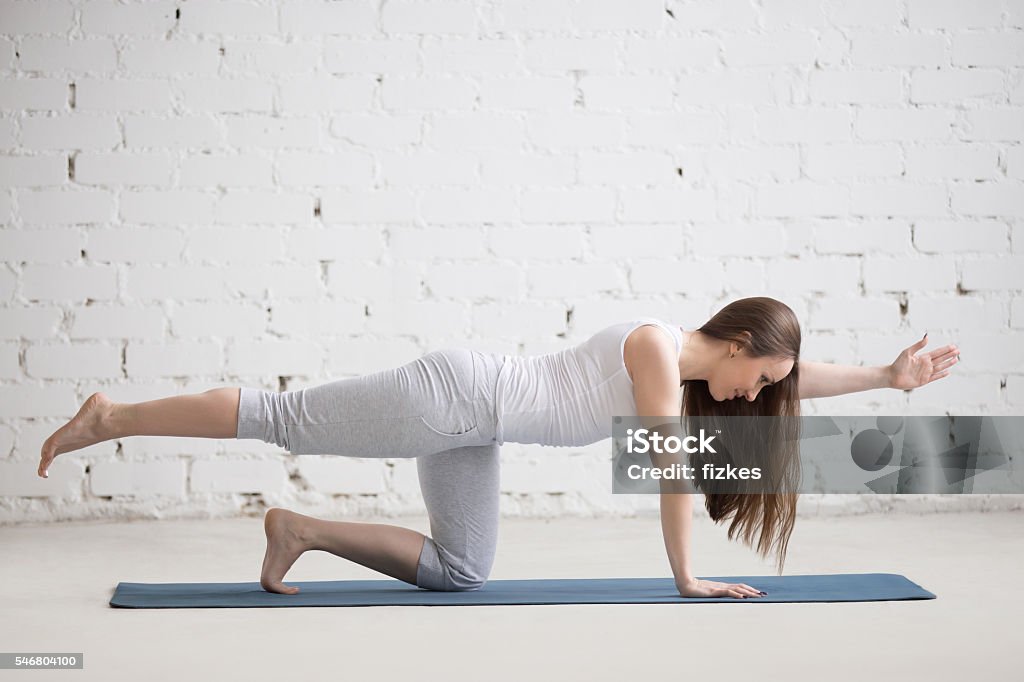 Portrait of beautiful woman doing bird-dog pose Attractive young woman working out indoors. Beautiful model doing exercises on blue mat in room with white walls. Bird-dog or kneeling opposite arm and leg extension (chakravakasana). Full length Exercising Stock Photo