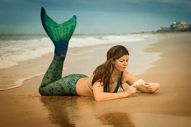 Caucasian woman in late 30's dressed up as a mermaid wearing a blue and teal green tail lies on her stomach in the sand and examines a shell at the Atlantic Ocean, Vilano Beach, St. Augustine, Florida, USA