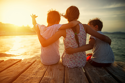 Brothers and sister sitting and embracing at sea pier. Summer vacations day evening.