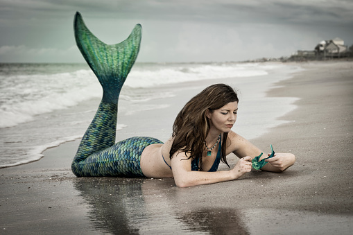 Caucasian woman in late 30's dressed up as a mermaid wearing a blue and teal green tail lies on her stomach in the sand and examines a turquoise colored sea horse at the Atlantic Ocean, Vilano Beach, St. Augustine, Florida, USA