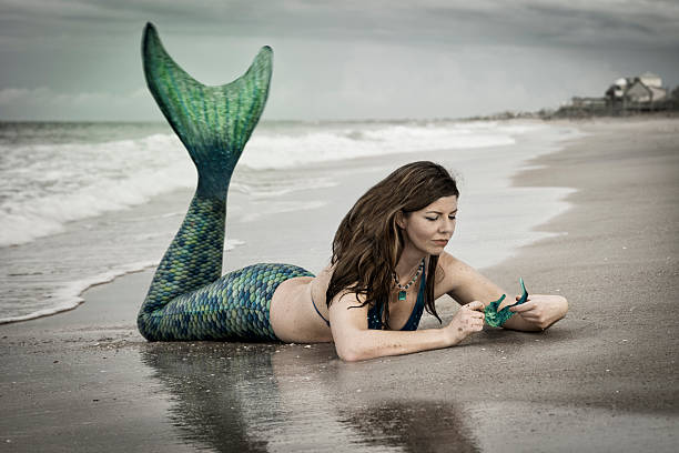 Fantasy mermaid with sea horse Caucasian woman in late 30's dressed up as a mermaid wearing a blue and teal green tail lies on her stomach in the sand and examines a turquoise colored sea horse at the Atlantic Ocean, Vilano Beach, St. Augustine, Florida, USA animal fin photos stock pictures, royalty-free photos & images