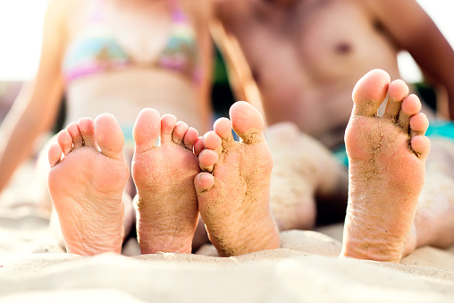 Feet of unrecognizable couple sitting on the sandy beach, sunny summer heat, barefoot