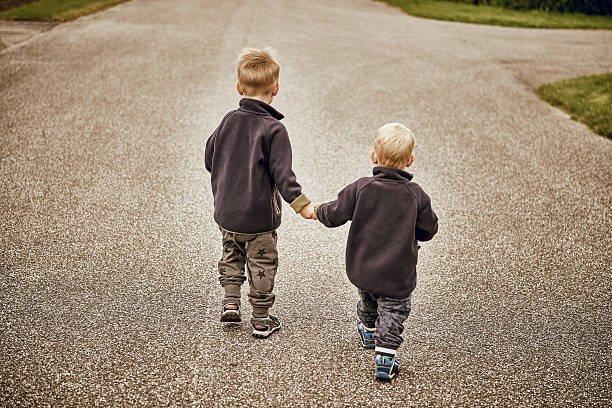 2 boys running hand in hand away from the camera in the street in a suburban area. The boys wear gray clothes. The composition is sparse.