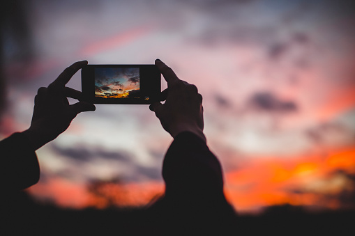 Over-shoulder view of a person taking a picture of a sunset on their smartphone.