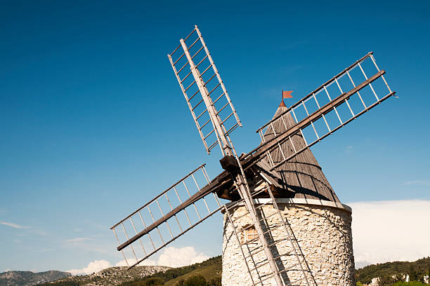 A historic stone windmill with wooden sails set against a vibrant blue sky and fluffy clouds, surrounded by lush green trees.