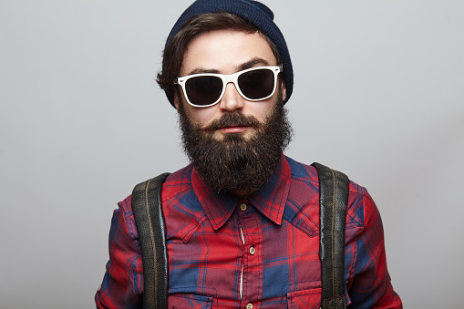 Portrait brutal bearded hipster man over grey background. Young man face closeup wearing a hat and backpack.