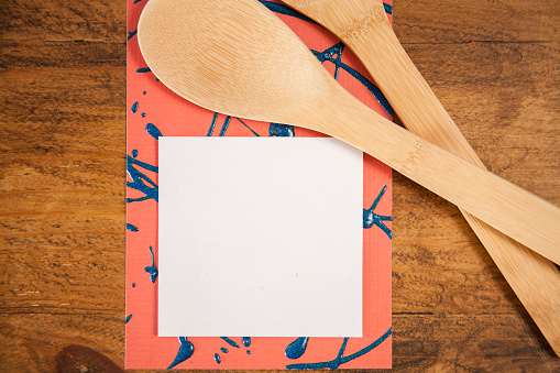 Wooden kitchen tools, spoon, spatula on wooden kitchen table with blank adhesive notepad for your copy.  Notepad on unique red and blue background.