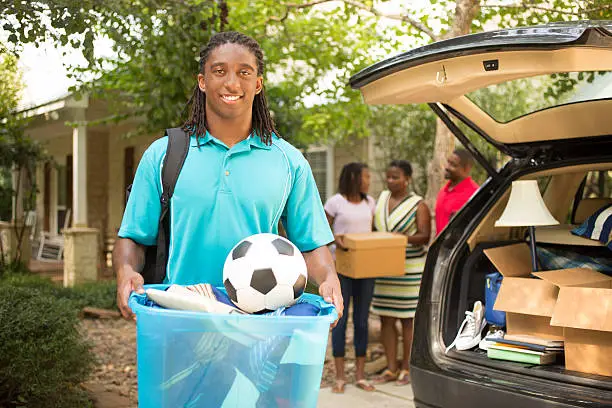 African descent boy heads off to college or moves away from home.  The 18-year-olds' parents and sister are all helping him pack up his car as he gets ready for the big move.  He is excited to start his college adventures. He wears a backpack and carries a box of belongings.  Family events.  Back to school.
