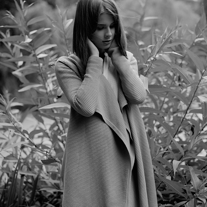 Black and white photo of a young woman in coat holding hands on her neck, nature on background.