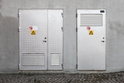 Metal doors with High Voltage warning signs in gray concrete industrial wall, background photo texture, front view