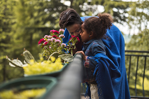 Young African American mother and daughter enjoying in a smell of flowers on a balcony.