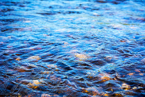 Defocused blue water surface background. Stony bottom of a clear mountain river. Flecks of sunlight and ripples on the water of river. Water background.  Water surface texture.