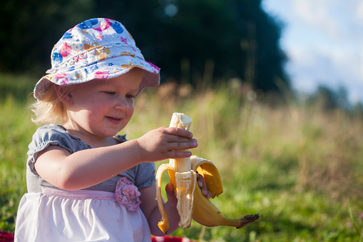 Little smiling girl is holding banana in her hand in sunny day, some copyspace on the right.