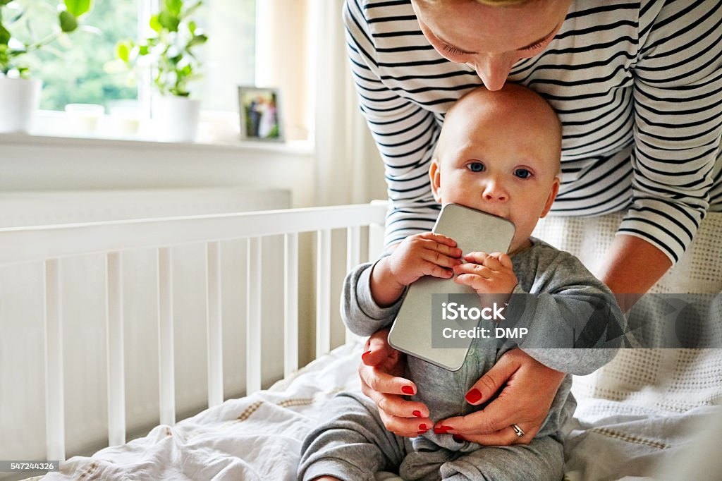 Baby boy chewing a cellphone in crib Cute baby boy in the crib chewing a cellphone with his mother by at home Baby - Human Age Stock Photo