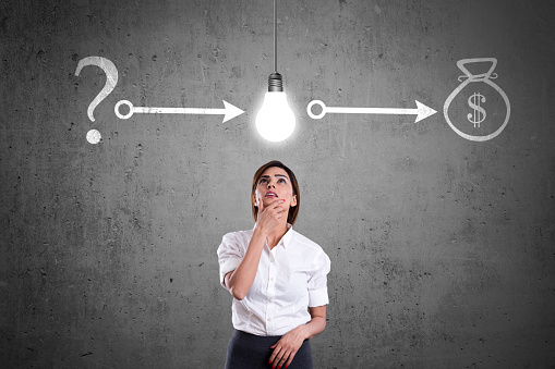 Young businesswoman standing with hands on chin and looking up at glowing light bulb.