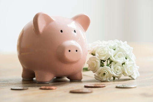 Loose coins are lying on a wooden table in front of a pink ceramic piggy bank. There is a small wedding bouquet of miniature white roses beside the moneybox on the table. The coins are to be deposited into the piggy bank to save for the wedding.