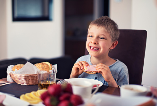 Cute happy little boy eating breakfast. The boy is aged 6 and is laughing happily holding a sandwich in his hands.