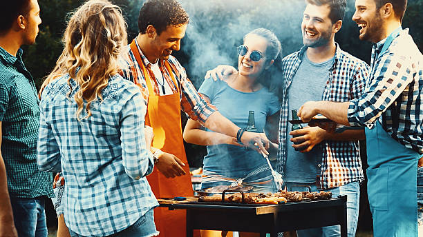 Barbecue party. Closeup side view of group of mid 20's people having backyard barbecue party. There are three guys and four girls gathered around heavily smoking grill and sipping cold beer. One of the guys is being today's chef. Toned image, mild contrast, back lit. barbecue grill photos stock pictures, royalty-free photos & images