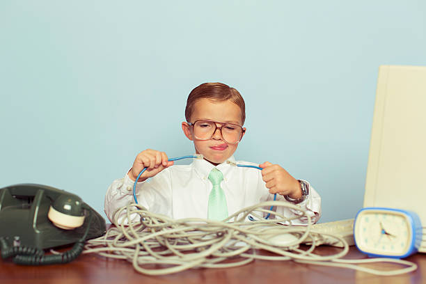 Young Boy IT Professional Smiles at Computer with Wire A young IT professional working with a large pile of tangled internet cables on his desk. He is dressed in a white shirt, tie and glasses while trying to reconnect his broken network. Retro styling. This IT technician can solve any network problem. nerd kid stock pictures, royalty-free photos & images