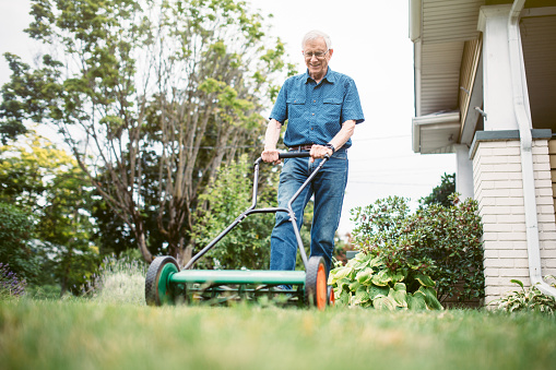 A man in his 70's does some mows the grass in his yard with a push lawnmower.  He has a content smile on his face.   A depiction of a relaxed and comfortable retirement.